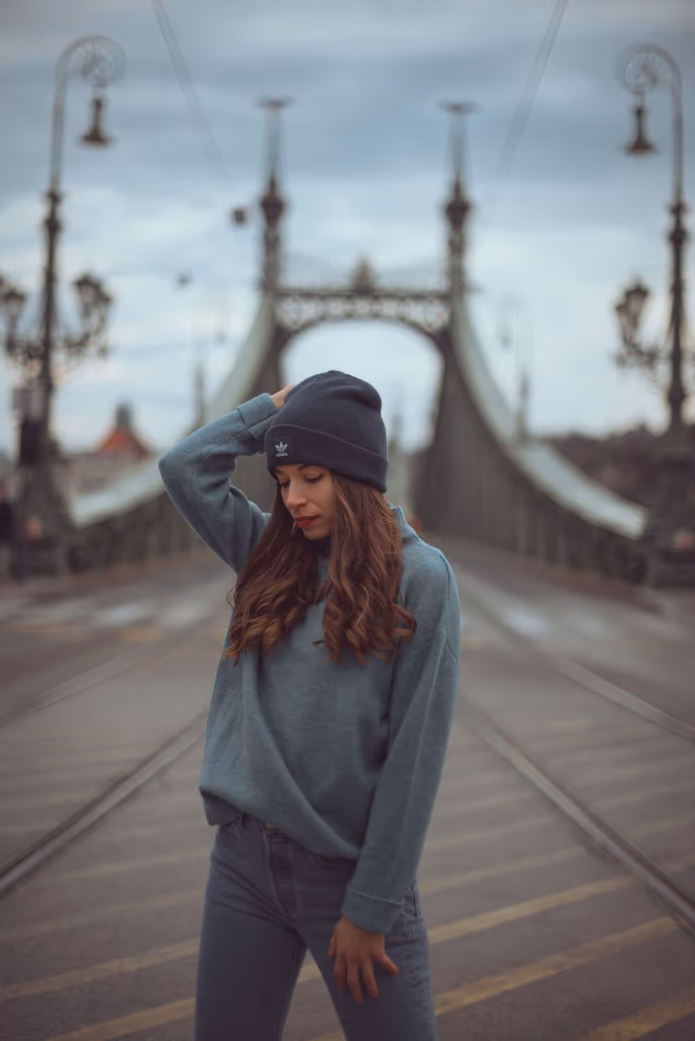 Young woman with long hair wearing a beanie and sweater standing on a bridge with vintage street lamps on a cloudy day.