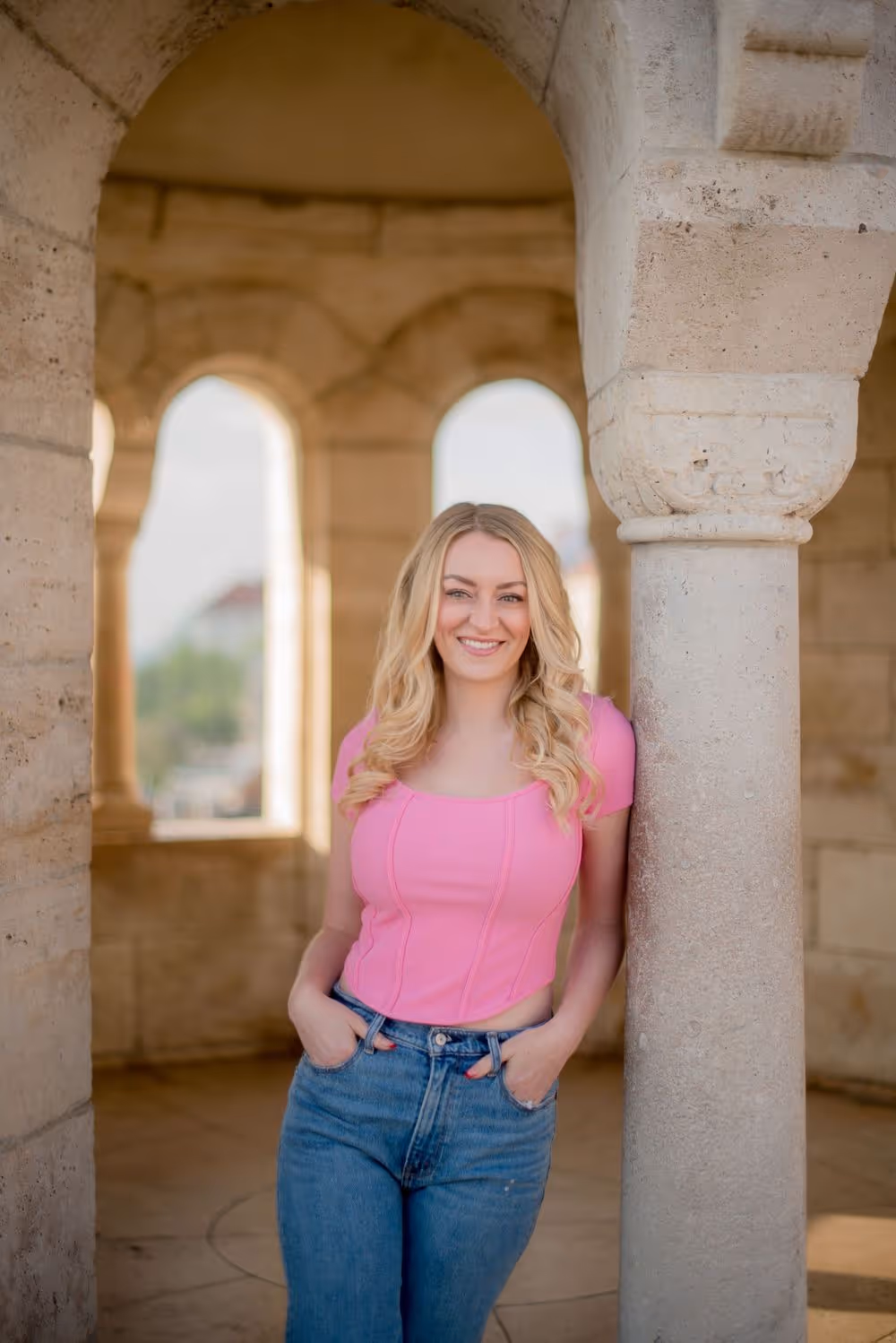 Smiling woman with blonde curly hair wearing a pink top and blue jeans leaning against a stone pillar in an arched stone structure.