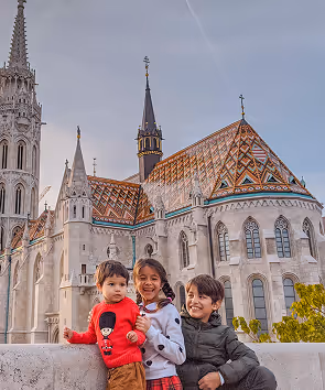Three children smiling and posing in front of a Gothic-style building with a colorful tiled roof.