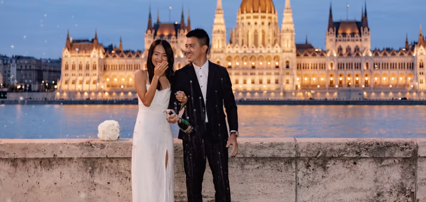 A couple dressed in formal attire popping a champagne bottle beside a river with a lit historic building in the background at dusk.