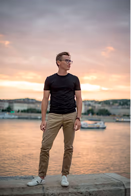 Young man in black t-shirt and khaki pants standing on a waterfront at sunset with buildings and a boat in the background.