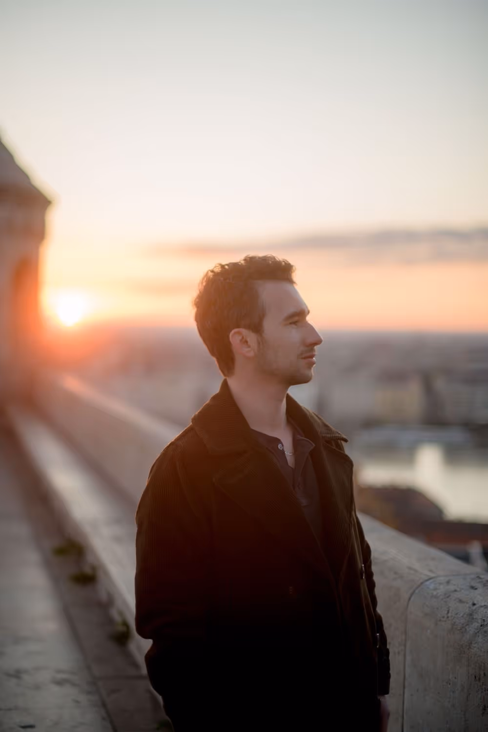 Man in dark coat standing outdoors near a stone railing at sunset with a blurred cityscape background.