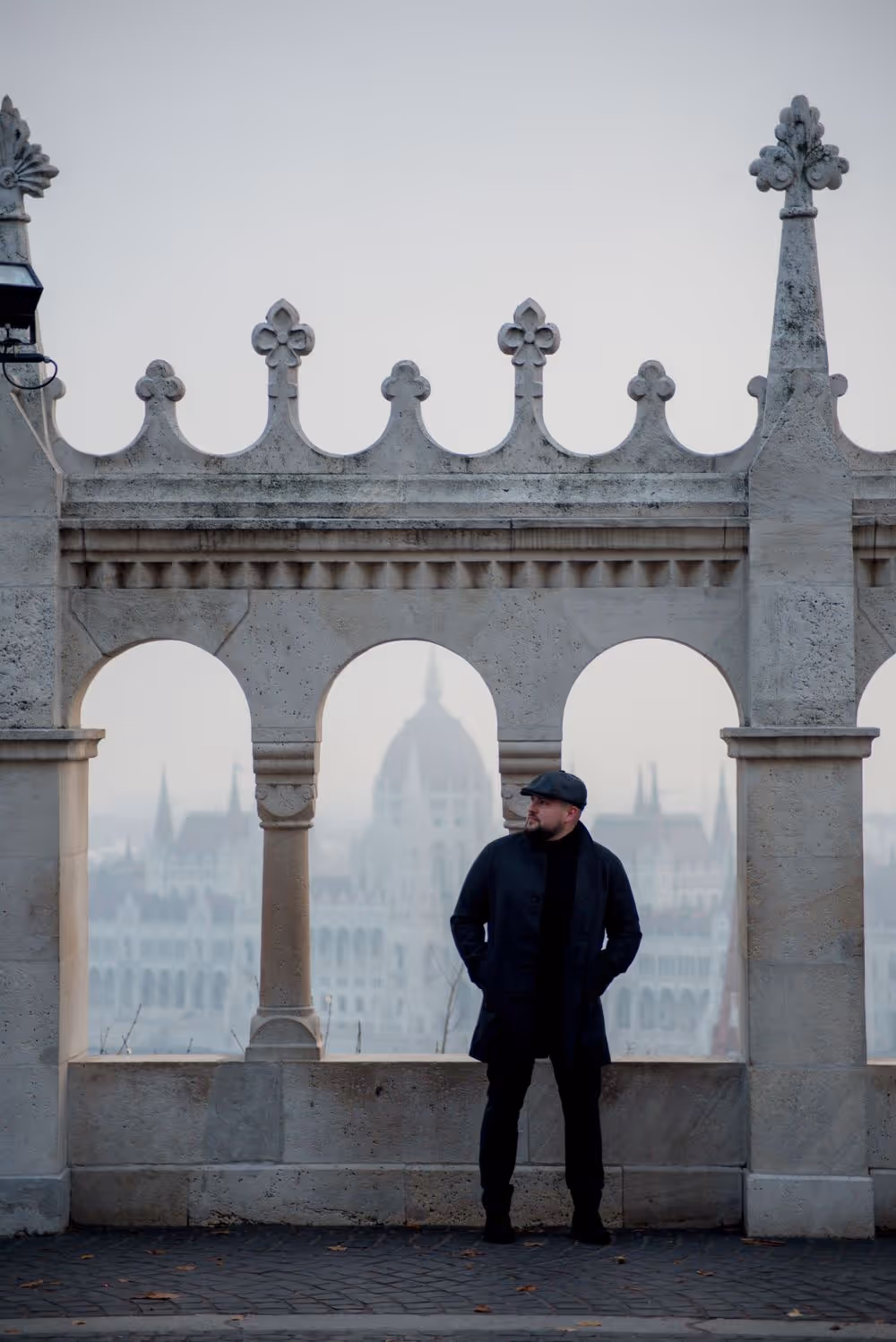 Man in a dark coat and cap standing under stone arches with a blurred cityscape in the background.