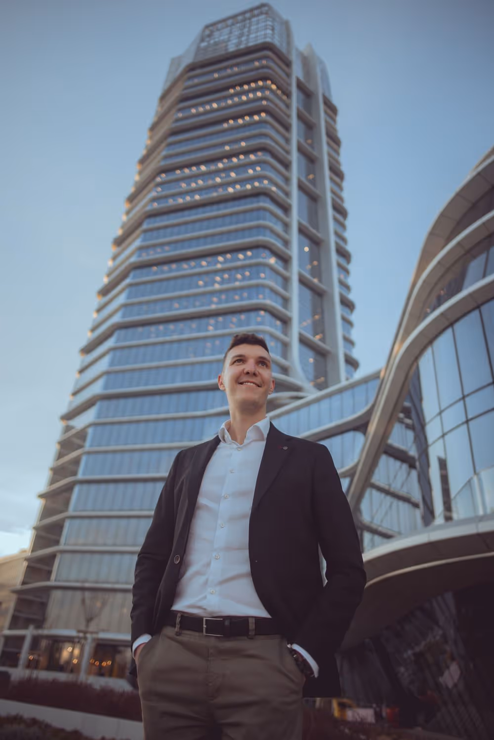 Young man in business casual attire standing confidently in front of a modern glass skyscraper.