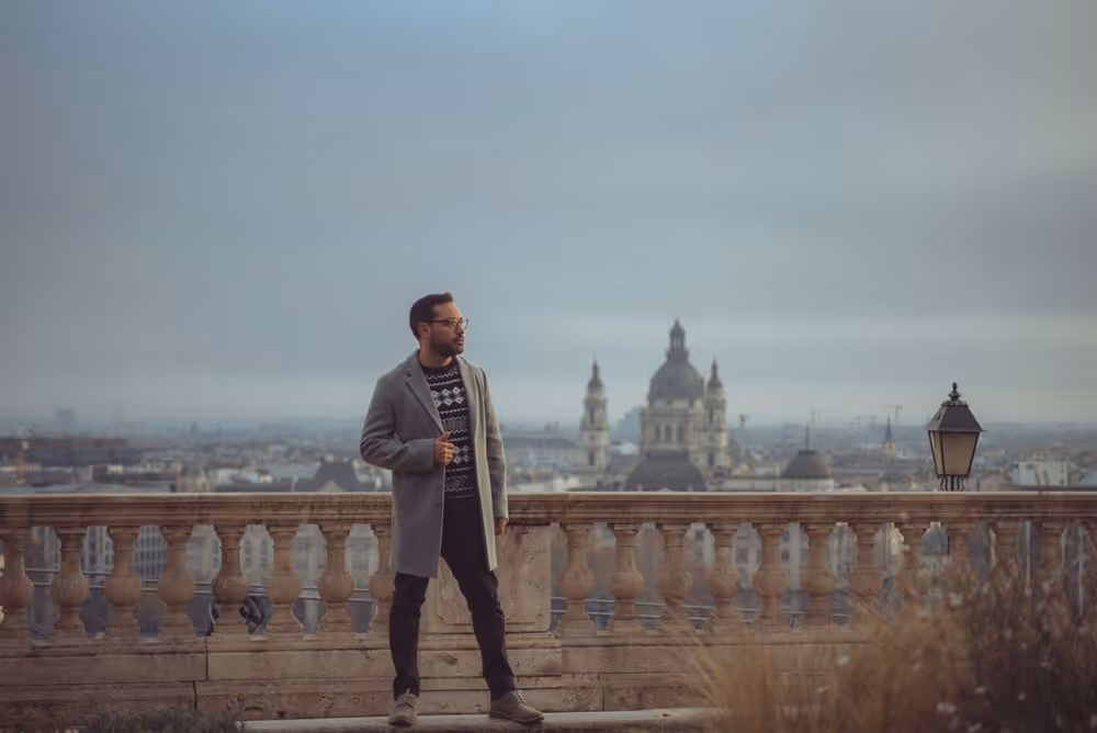 Man wearing a gray coat and patterned sweater standing on a stone terrace overlooking a city with historic buildings under an overcast sky.