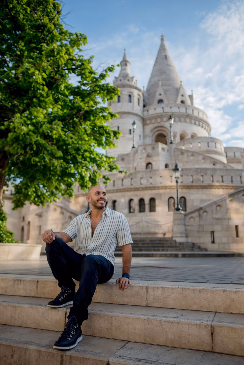 Man sitting on stone steps with a historic castle featuring towers and arches in the background under a partly cloudy sky.