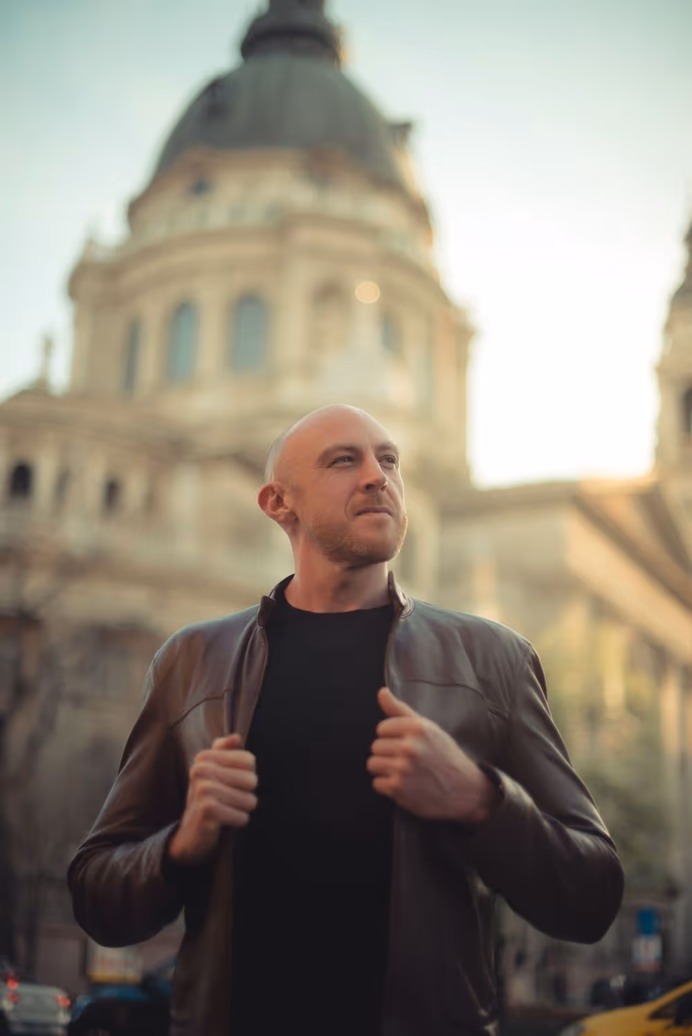 Bald man wearing a leather jacket stands confidently in front of a historic building with a large dome.