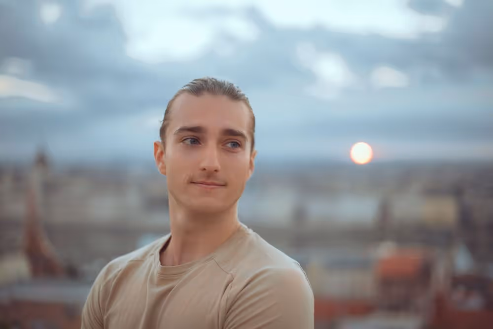 Young man with hair tied back wearing a beige shirt, looking to the side with a blurred cityscape and sunset in the background.