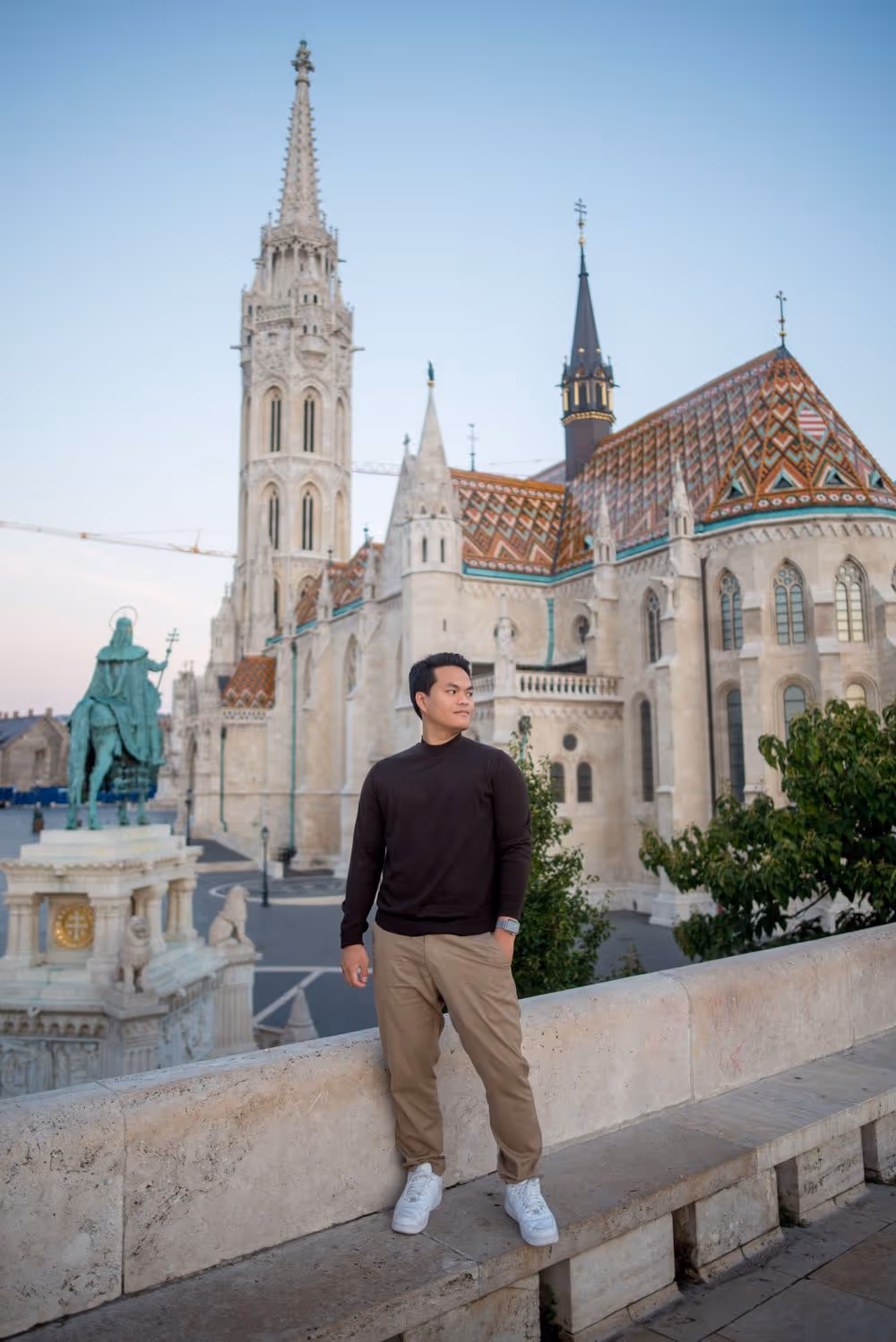 Man in black sweater and beige pants standing on a stone ledge with historic gothic-style building and equestrian statue in the background.