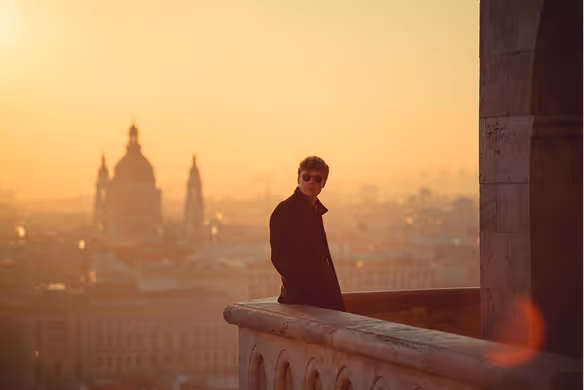 Person in dark clothing and sunglasses leaning on a stone railing overlooking a city at sunset with a blurred cathedral in the background.