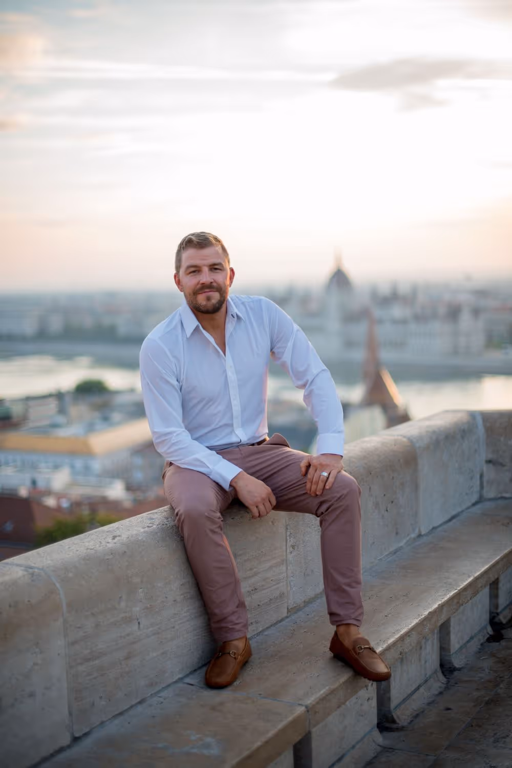 Man in a white shirt and brown pants sitting on a stone bench overlooking a cityscape at sunset.