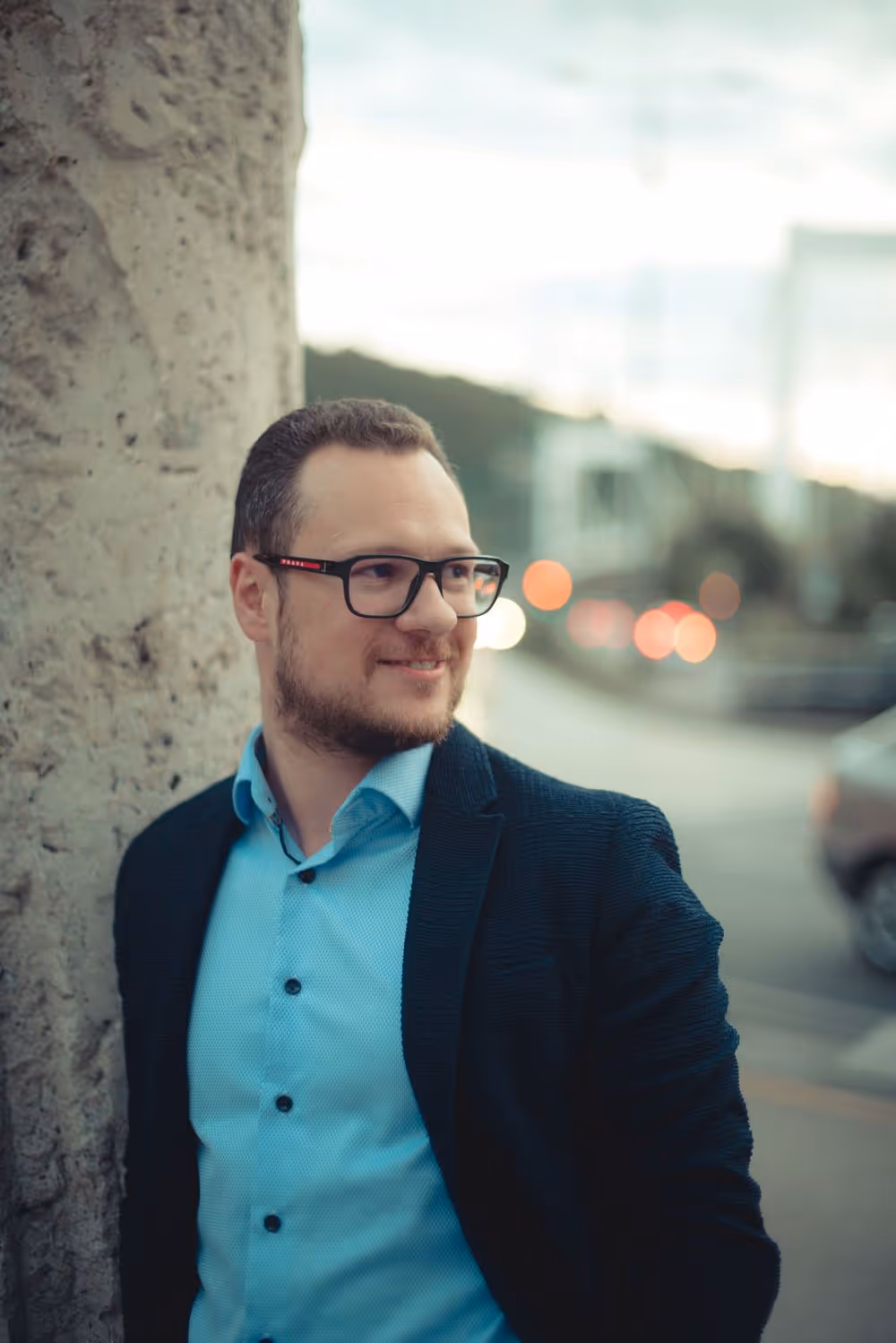 Man wearing glasses and a blue shirt with a dark jacket, leaning against a textured wall, smiling and looking to the right with blurred city lights in the background.