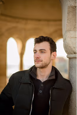 Young man in a black jacket leaning against a stone pillar under arches, looking thoughtfully to the side.