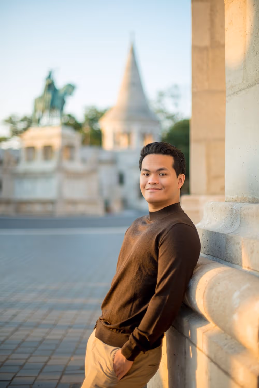 Young man in brown sweater leaning against stone wall outdoors with blurred historic monument in the background.