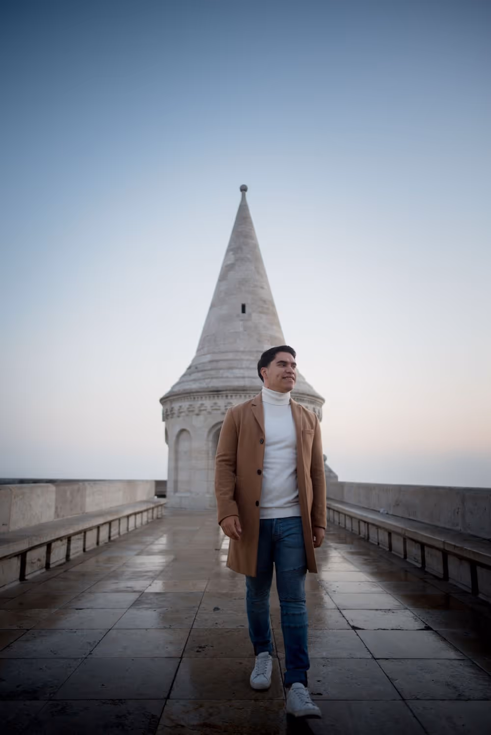 Man in white turtleneck, brown coat, and jeans walking on tiled pavement with a tall stone tower in the background under a clear sky.