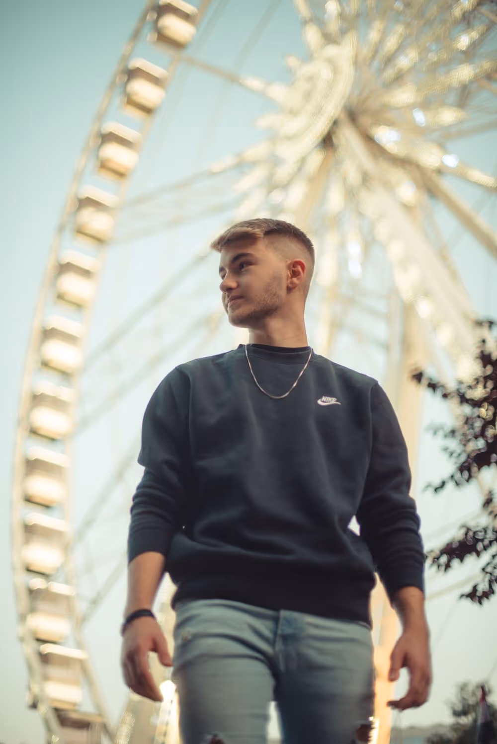 Young man wearing a black Nike sweatshirt and light blue jeans standing in front of a large Ferris wheel.