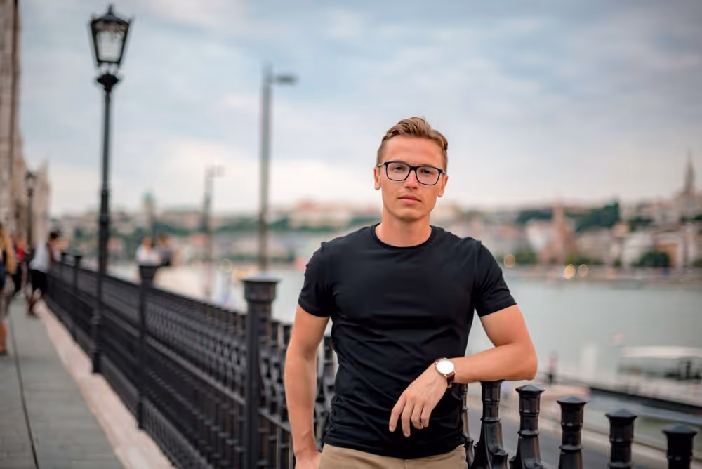Young man wearing glasses and a black t-shirt leaning on a black metal railing by a river with a cityscape in the background.