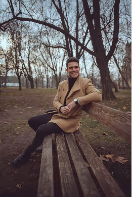 Smiling man in a camel coat sitting on a wooden bench in a park with bare trees.