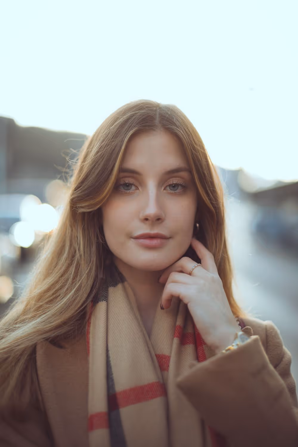 Close-up of a young woman with long light brown hair wearing a beige scarf and brown coat, gently touching her face.