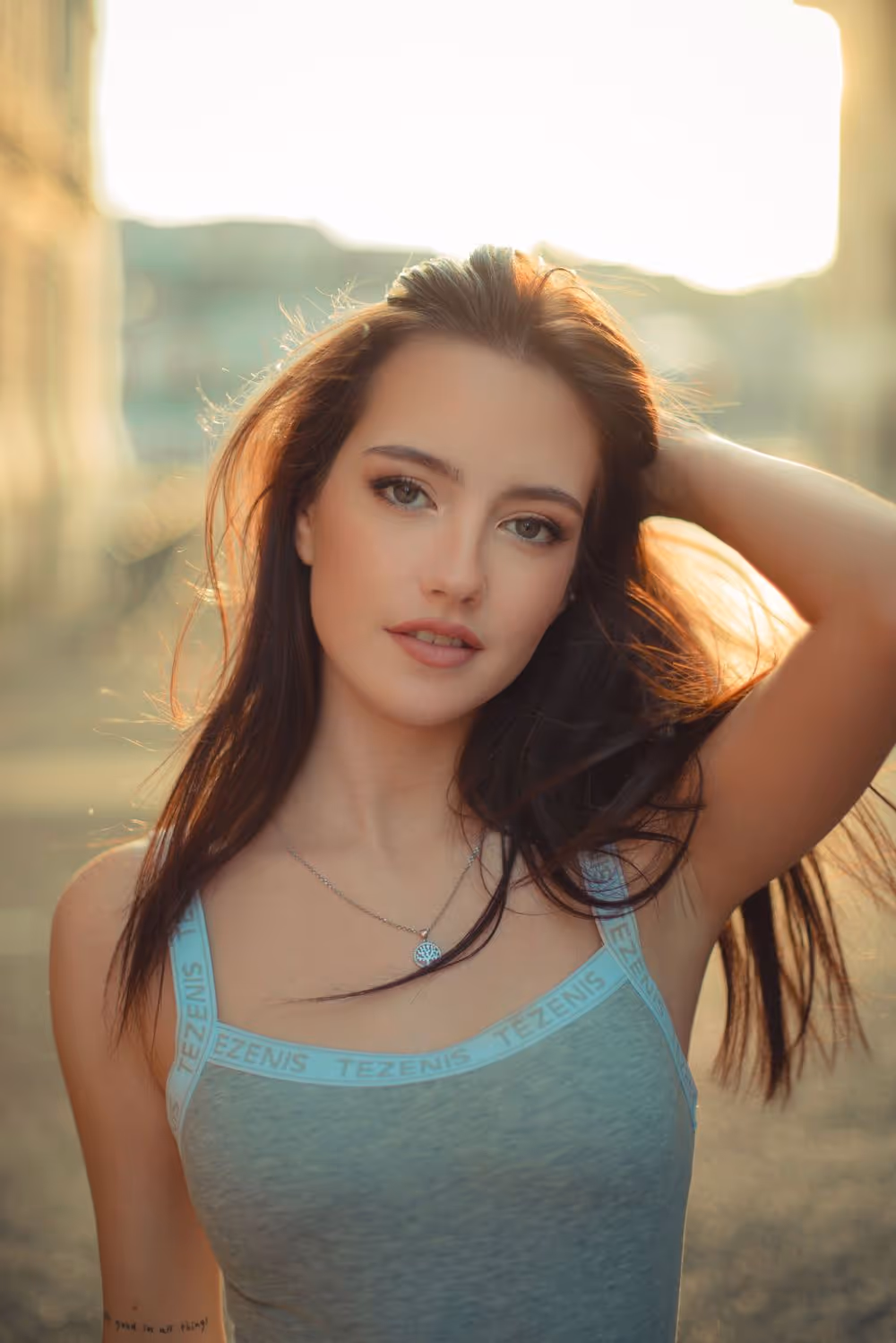 Young woman with long brown hair wearing a gray tank top and necklace, posing with one hand behind her head in warm sunlight.