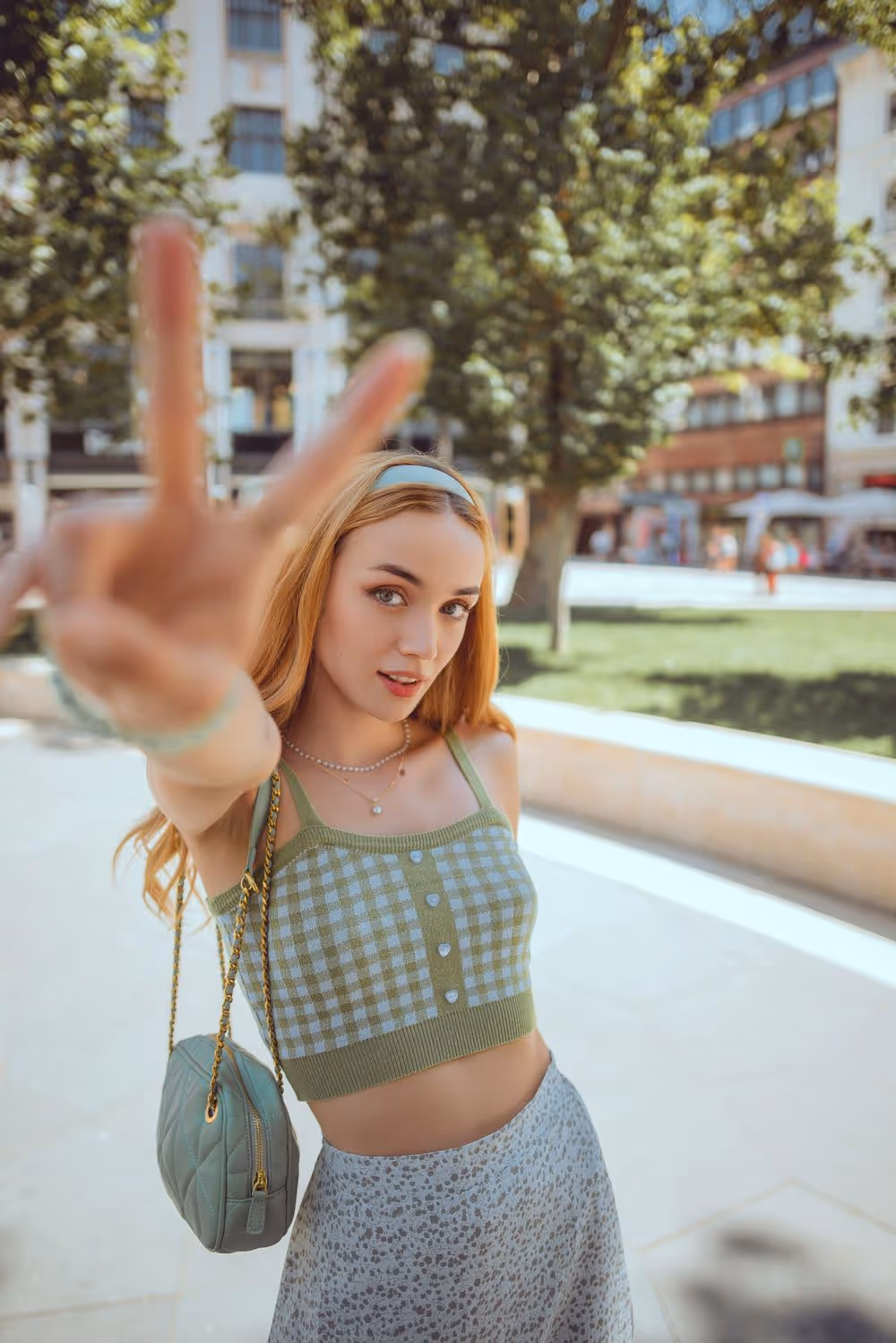 Young woman outdoors wearing a green checkered crop top and floral skirt, making a peace sign with her hand toward the camera.