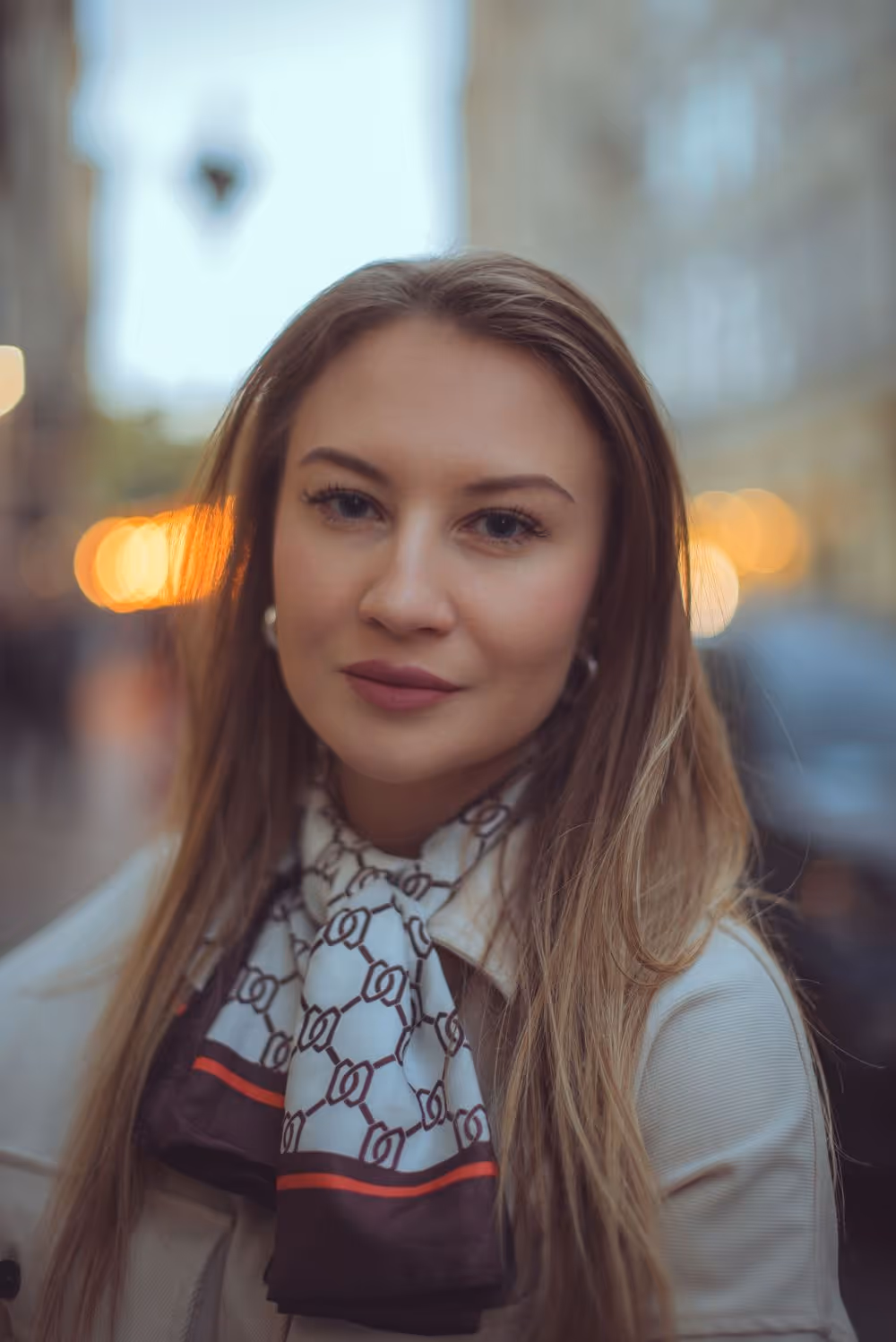 Young woman with long blonde hair wearing a white jacket and a patterned scarf, standing outdoors with blurred city lights in the background.