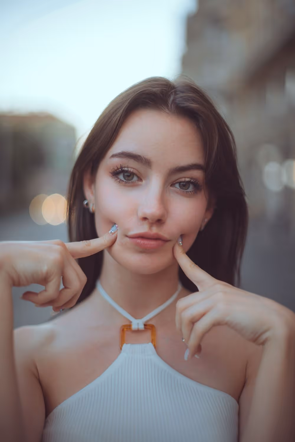 Young woman with long dark hair wearing a white halter top and touching her cheeks with both index fingers.