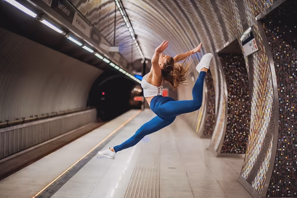 Female dancer in blue leggings and white top performing a high leap in an empty subway station.