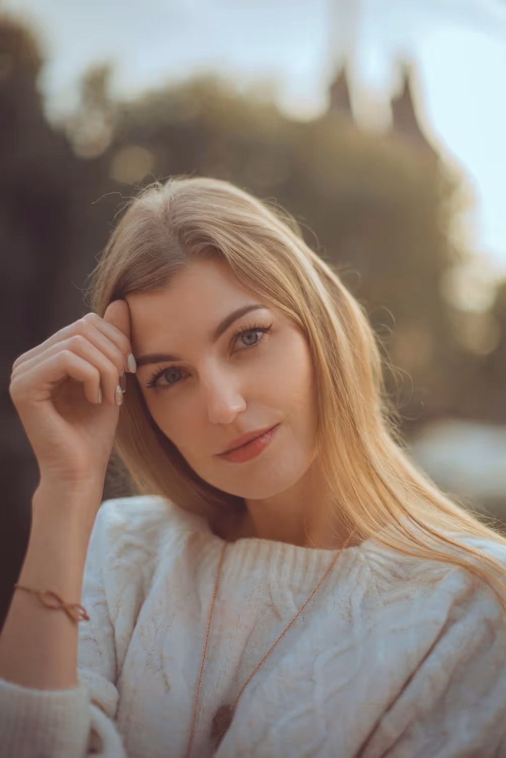 Young woman with long blonde hair wearing a white knit sweater, resting her head on her hand and looking softly at the camera.