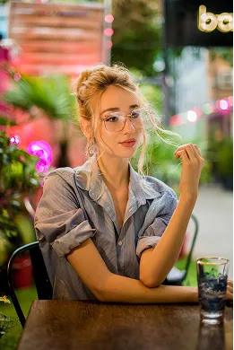 Young woman with glasses and a gray shirt sitting at a table in a colorful outdoor setting, looking thoughtfully to the side.
