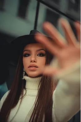 Young woman with long dark hair wearing a black hat and white turtleneck sweater reaching hand towards the camera.