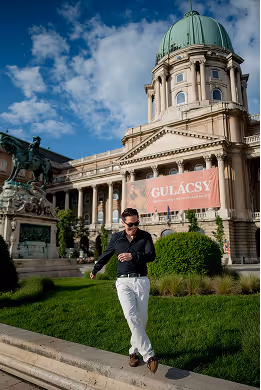 Man in sunglasses and black shirt walking on a stone ledge in front of a historic building with a green dome and a statue of a horse and rider.