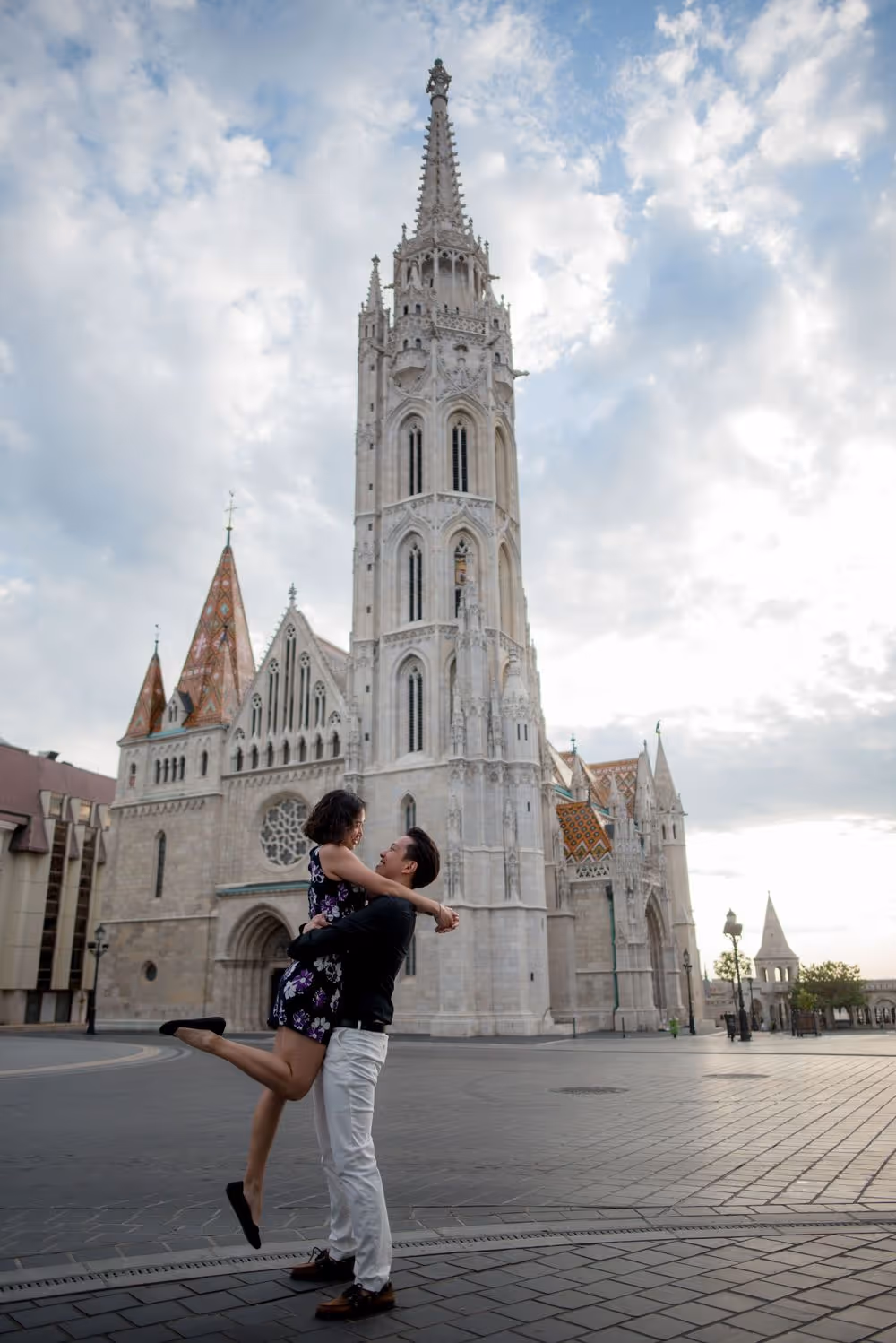 Couple embracing joyfully near a historic cathedral with a tall spire under a cloudy sky.