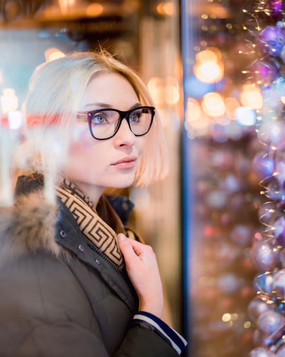 Young woman with glasses and blonde hair looks thoughtfully at a festive display of purple and silver Christmas ornaments through a window.