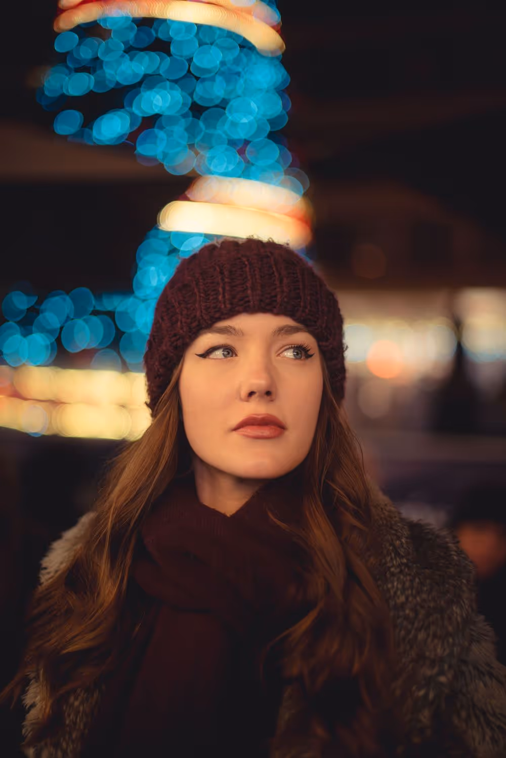 Woman wearing a knit hat and scarf standing outdoors at night with blurred blue and yellow lights in the background.
