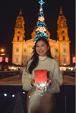 Smiling woman in white sweater holding a glowing red gift box with a decorated Christmas tree and illuminated historic building in the background at night.