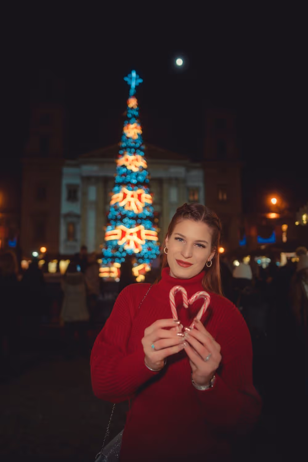 Woman in red sweater holding two candy canes forming a heart with a decorated Christmas tree and building lit behind her at night.