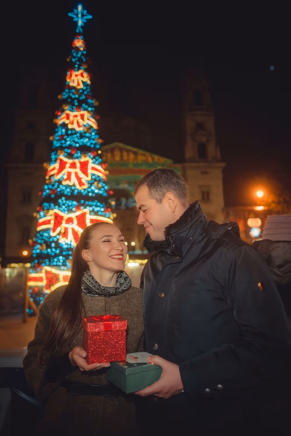 Smiling couple exchanging gifts in front of a decorated Christmas tree at night.