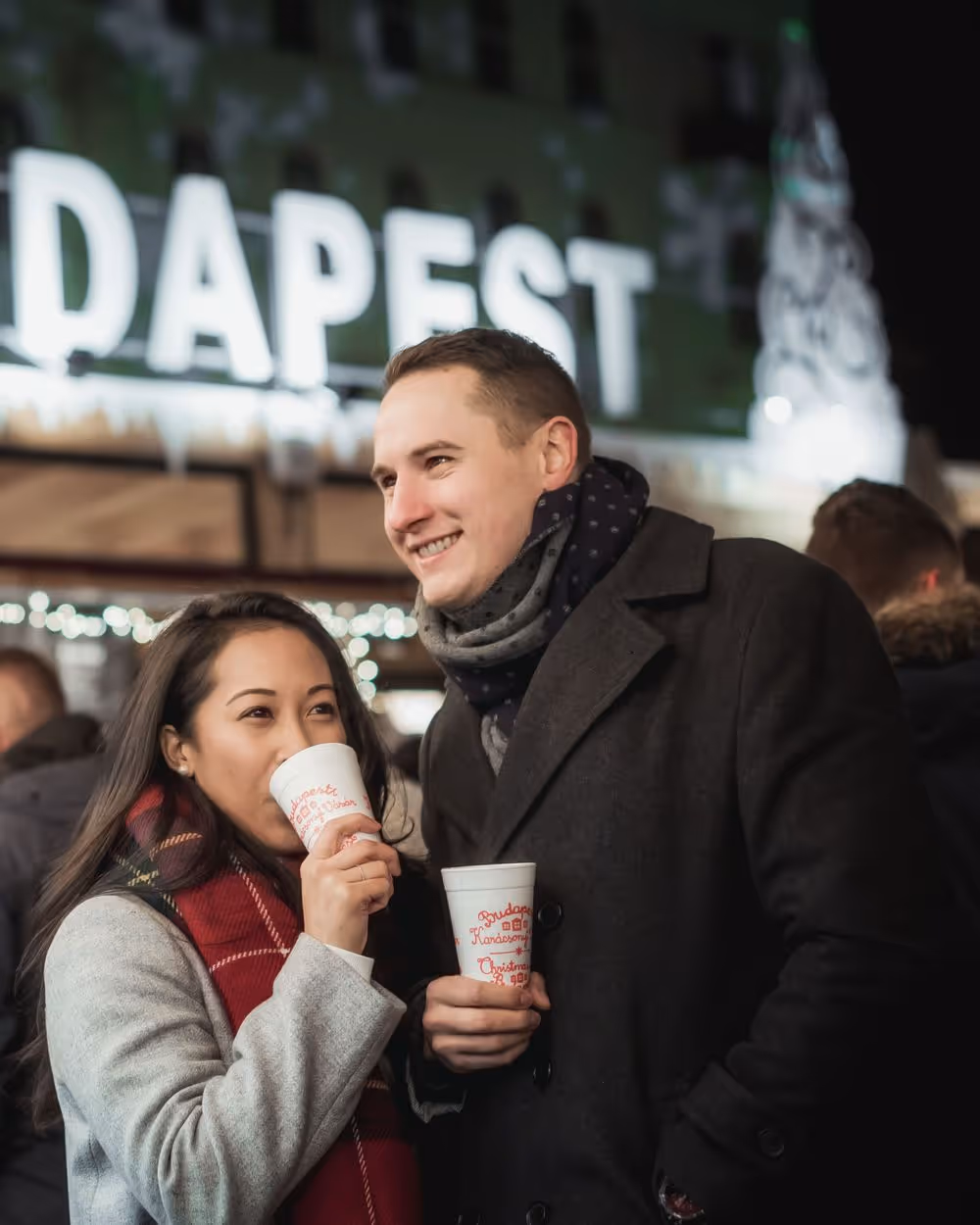 Smiling couple in winter coats drinking from festive cups at a night market with Budapest sign in background.