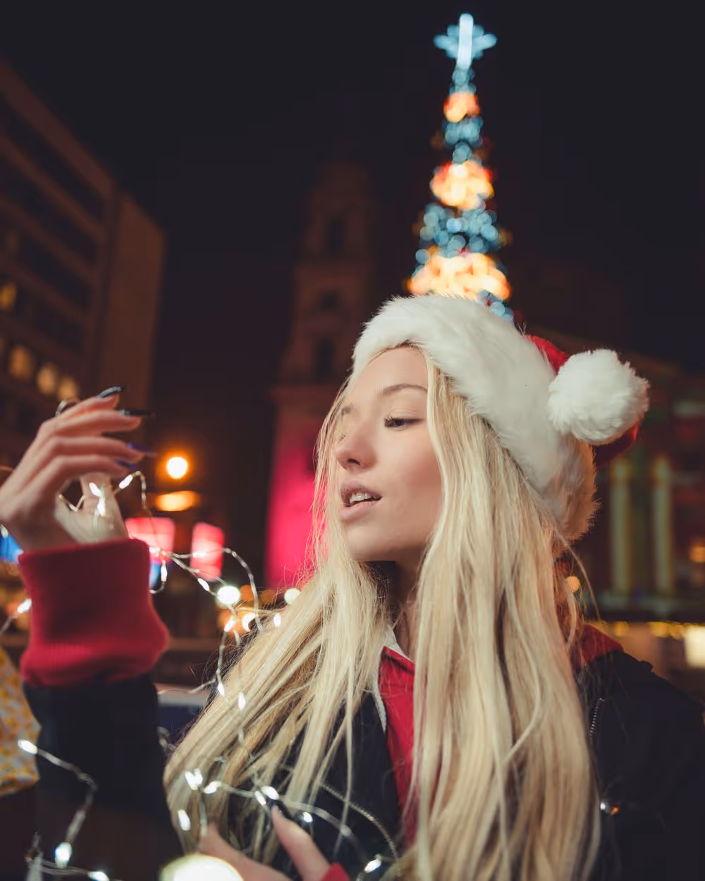 Young woman in a Santa hat holding and looking at glowing Christmas string lights at night, with a blurred lit Christmas tree in the background.