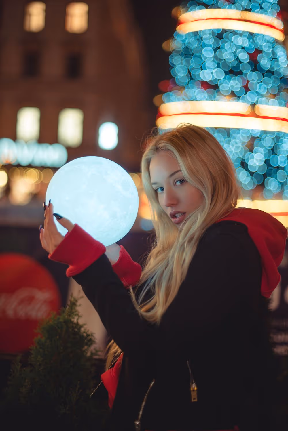 Young woman with long blonde hair holding a glowing sphere against a background of colorful holiday lights.