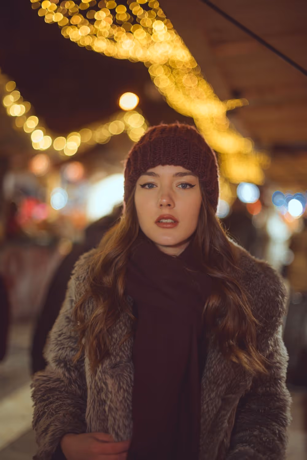 Young woman wearing a maroon knit hat and fur coat standing in a warmly lit outdoor evening setting with blurred string lights in the background.