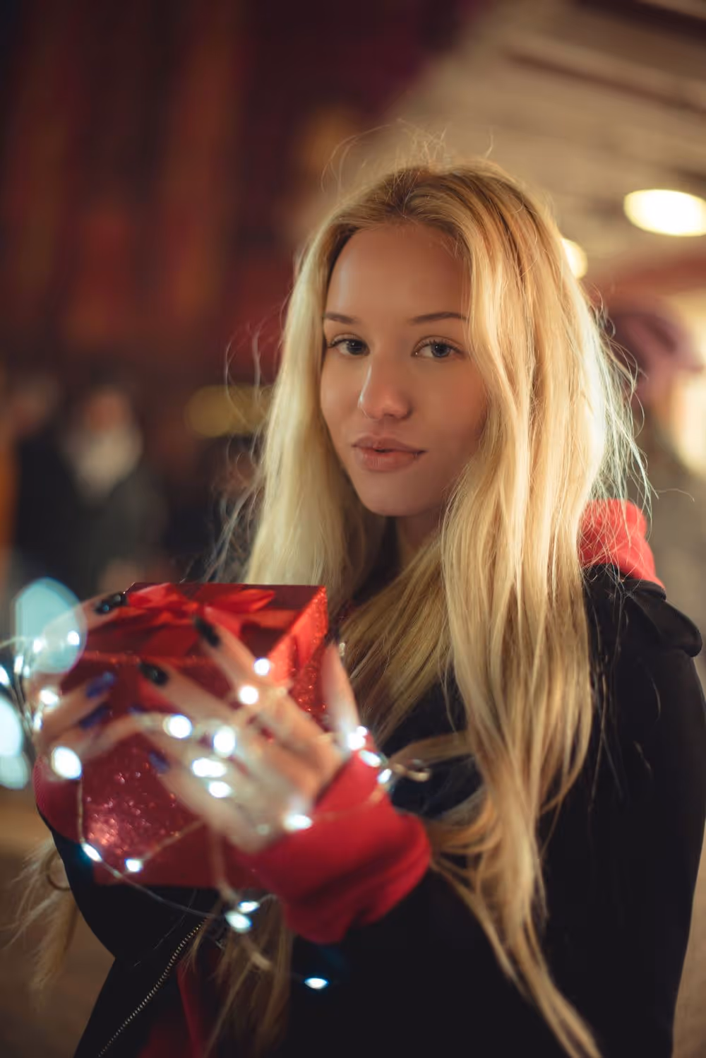 Blonde woman holding a red gift box wrapped with a bow, decorated with string lights.