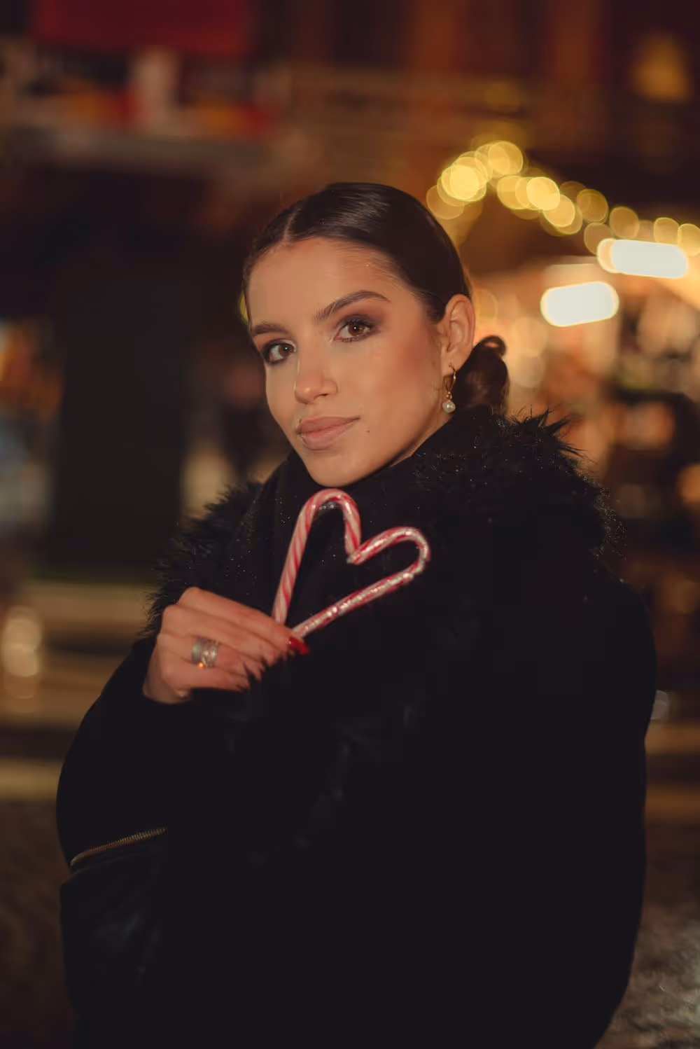 Woman wearing black coat holding two candy canes shaped like a heart with blurred warm lights in the background.